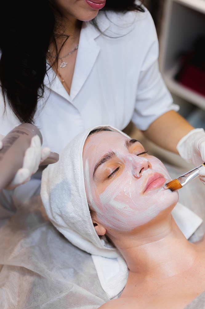 Beautician with a brush applies a white moisturizing mask to the face of a young girl client in a spa beauty salon.