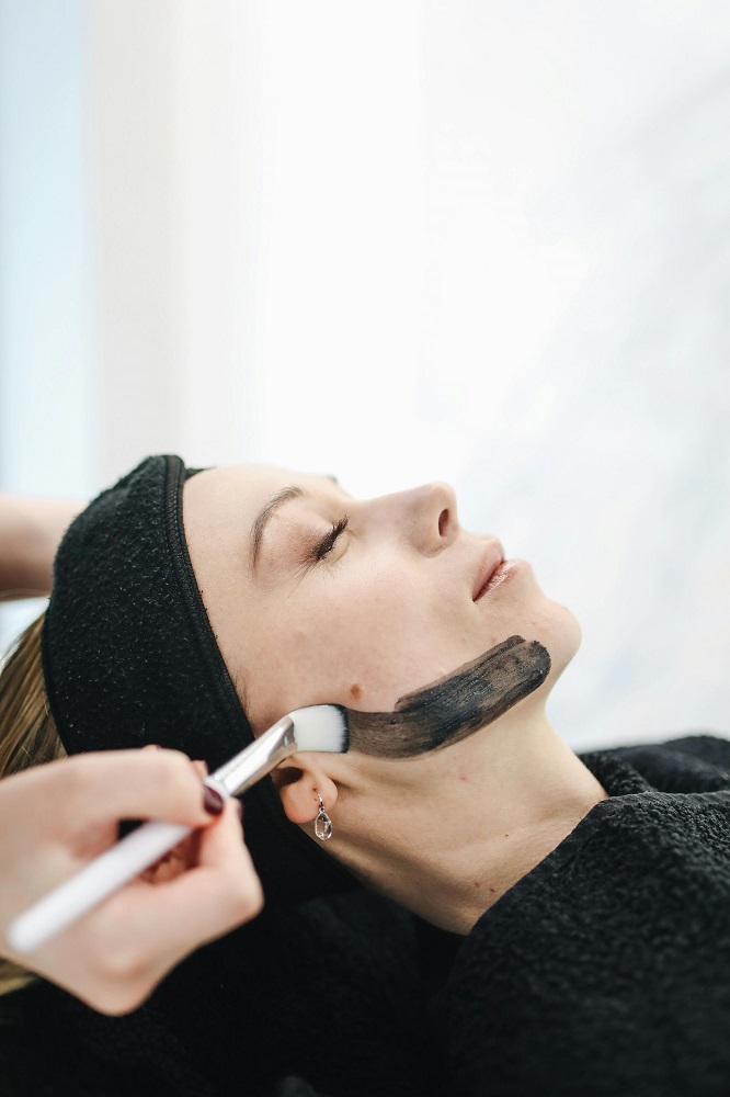 Beautician with a brush applies a black mask to the face of a female client in a spa beauty salon.