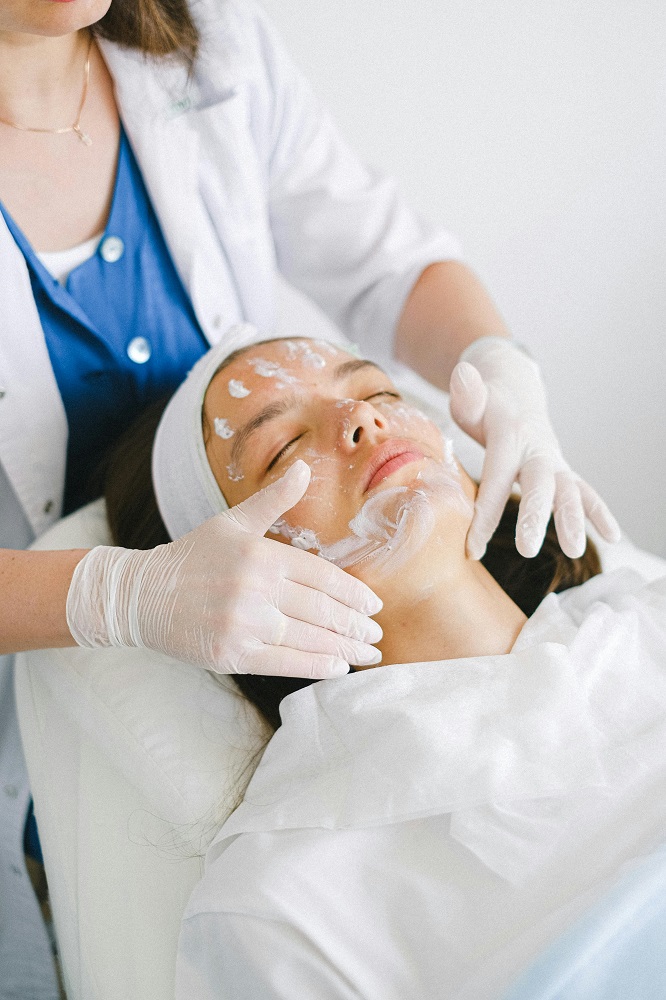 Beautician with a brush applies a white moisturizing mask to the face of a young girl client in a spa beauty salon.